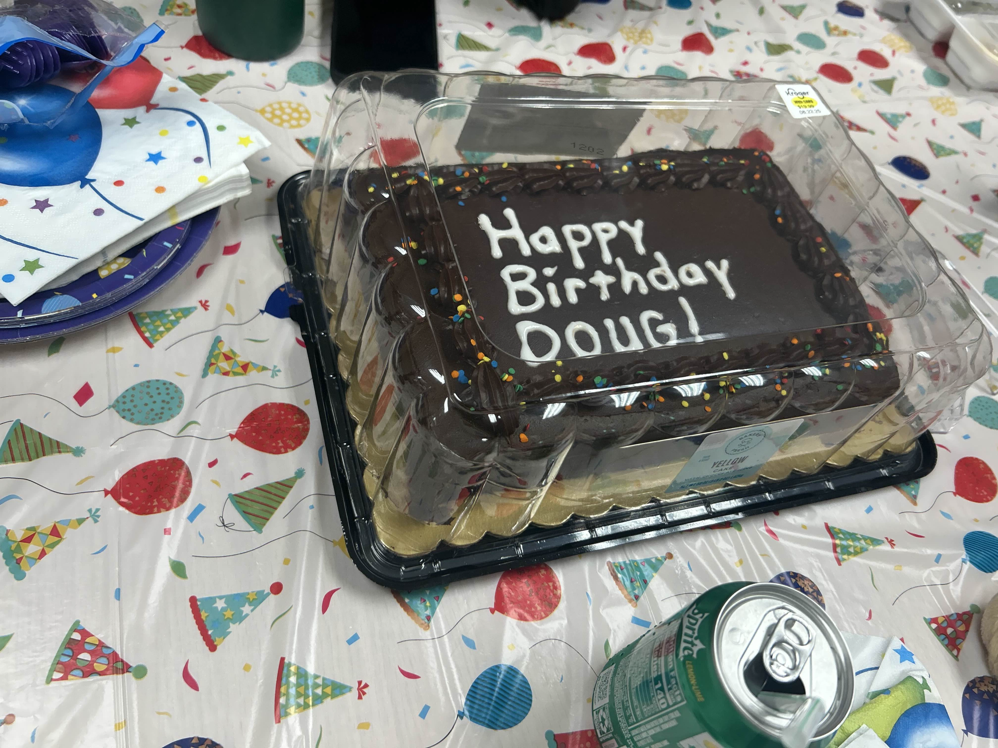 Image of a birthday cake sitting on a table with a plastic tablecloth on it. The cake has chocolate frosting and reads in white icing 'Happy Birthday Doug!'. There are drinks, plates, and other objects scattered on the table.